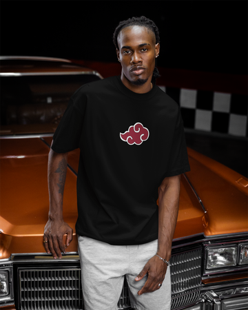 A man wearing a black short-sleeve t-shirt with a red and white Akatsuki cloud logo on the chest, paired with light gray shorts, standing in front of an orange classic car.